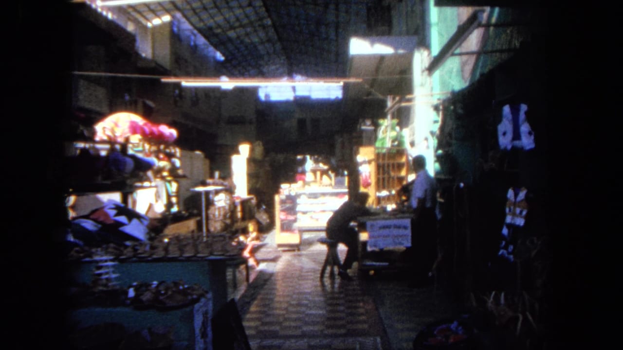 A seated adult appears at a counter inside the 1962 Tijuana market, with merchandise displays and bright lighting down the aisle.