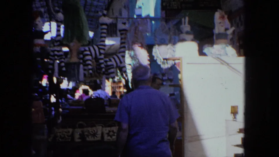 A rear view of the man walking below hanging textiles and patterned goods in the Tijuana market, with shoppers farther down the aisle.