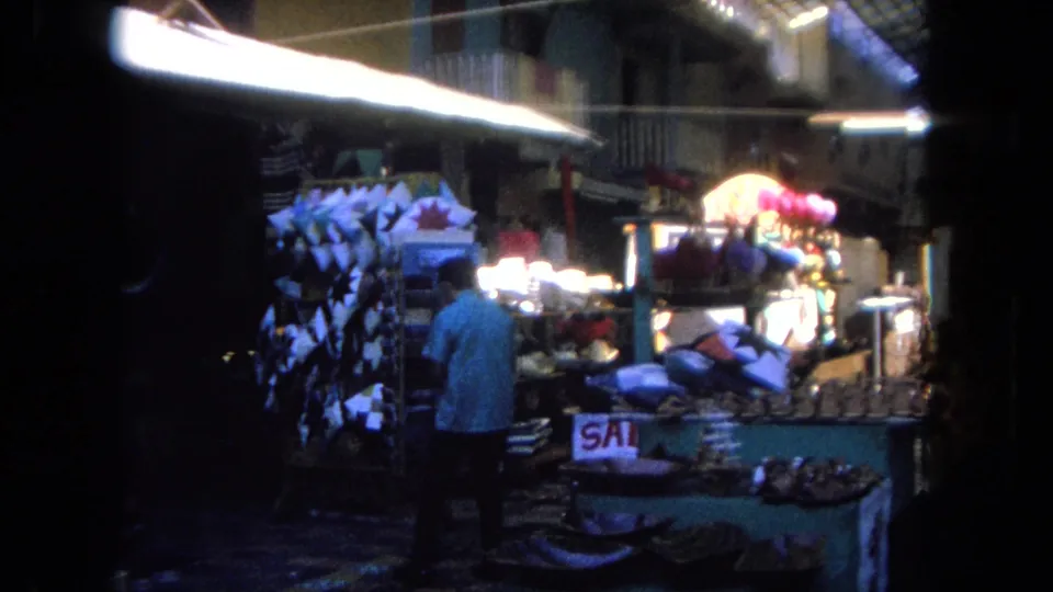 A Tijuana market stall displays hanging goods, textiles or pillows, overhead lighting, and a partially visible sale sign in the 1962 footage.