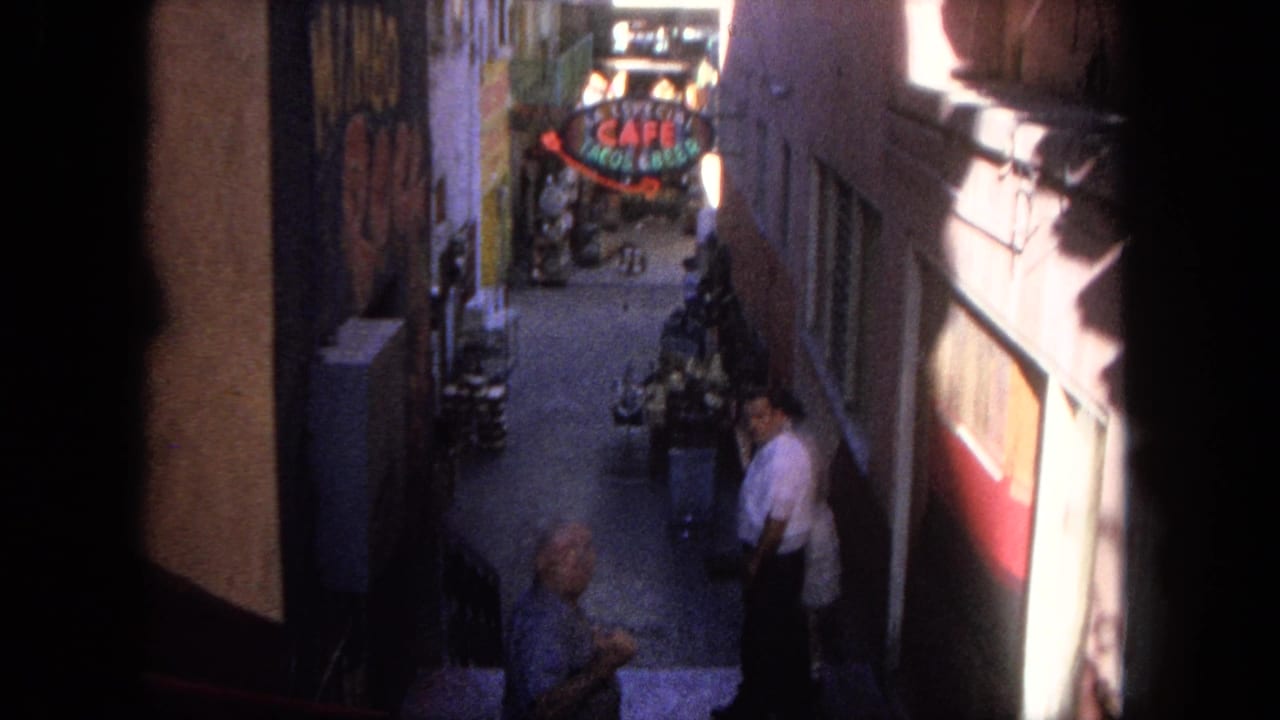 High-angle view down a narrow alley with pedestrians, painted walls, storefronts, and a cafe sign in the distance.