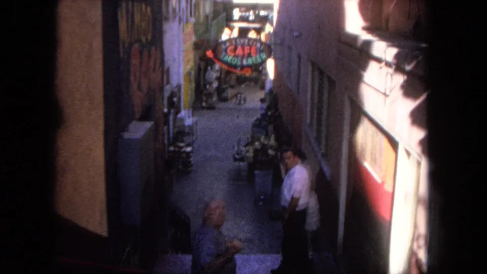 1962 Tijuana home-movie frame looking down a narrow commercial alley with pedestrians, strong shadows, painted walls, and a cafe sign.