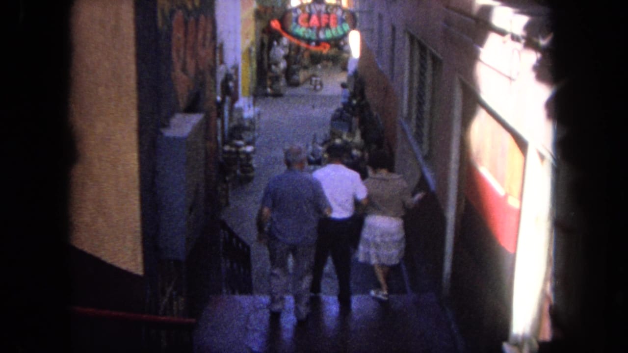 Three people walk away from the camera down a narrow alley toward a bright cafe sign.