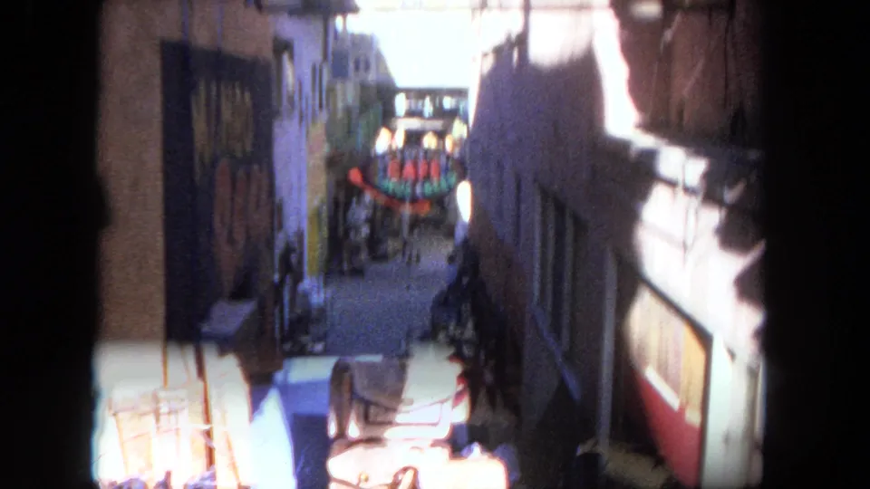 A 1962 Tijuana alley frame shows harsh sunlight near the foreground, painted walls, storefront edges, and the cafe sign in the distance.