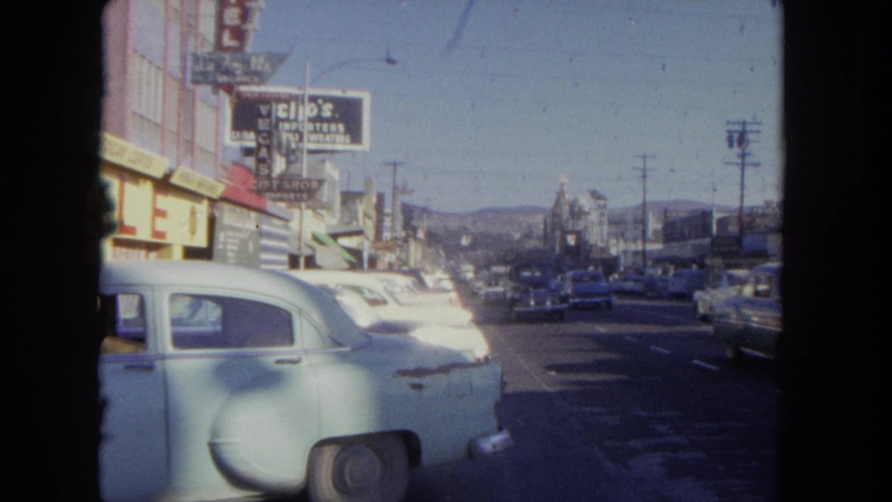 Downtown street with storefront signs, parked cars, and distant hills beyond the road.