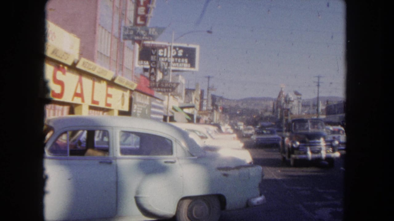 Street scene with a pale sedan near storefronts and traffic moving along the road.