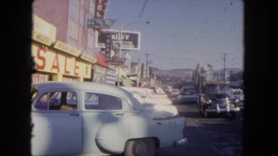 1962 Tijuana commercial street with a pale sedan in the foreground, shop signs, a SALE banner, and passing cars.