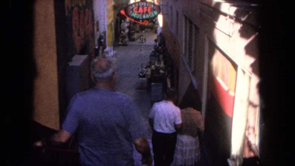 1962 Tijuana alley view with pedestrians on steps, close building walls, and a neon Cafe Tacos & Beer sign ahead.