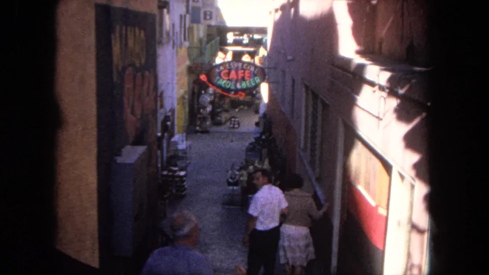 1962 Tijuana alley scene with pedestrians, tight building walls, shaded walkway, and a neon Cafe Tacos & Beer sign.