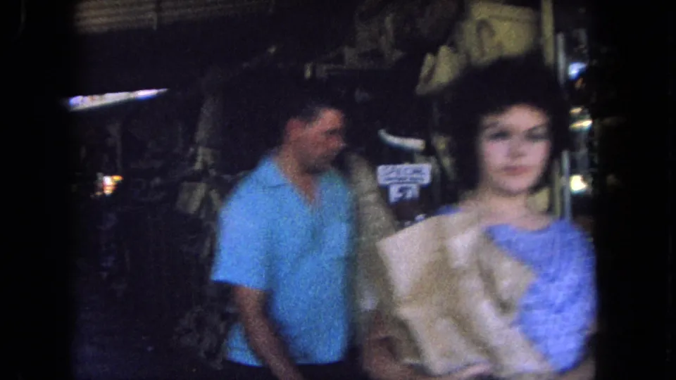 A close, softly focused 1962 market shot shows the couple moving past the camera as the woman holds a wrapped purchase.