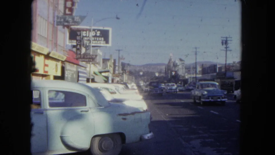 A sunlit Tijuana street scene shows mid-century cars, utility poles, storefronts, and visible commercial signs along the road.
