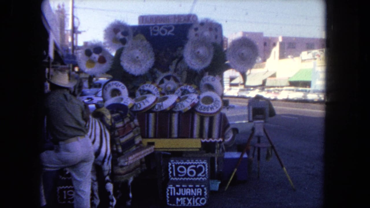 1962 Tijuana street scene with a man beside a souvenir cart covered in sombreros, textiles, and a zebra-patterned figure near traffic.