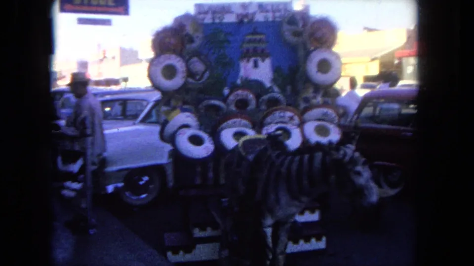 Tijuana street vendor cart viewed from behind, with sombreros, a painted panel, parked cars, and pedestrians on the sidewalk.