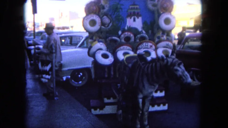 1962 Tijuana street view with a decorated souvenir cart, sombreros, zebra-patterned figure, pedestrians, and cars in the background.