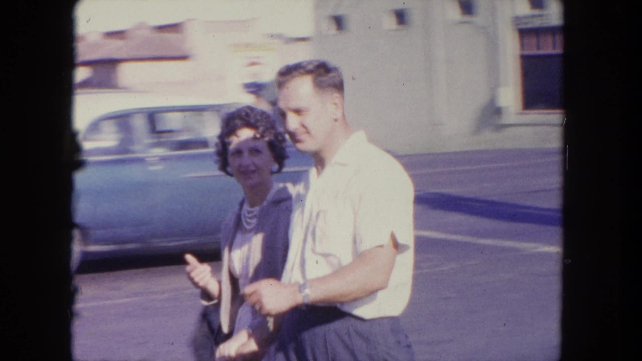 In Tijuana, Mexico, a woman and man walk past the camera on a sunny street with mid-century cars behind them.