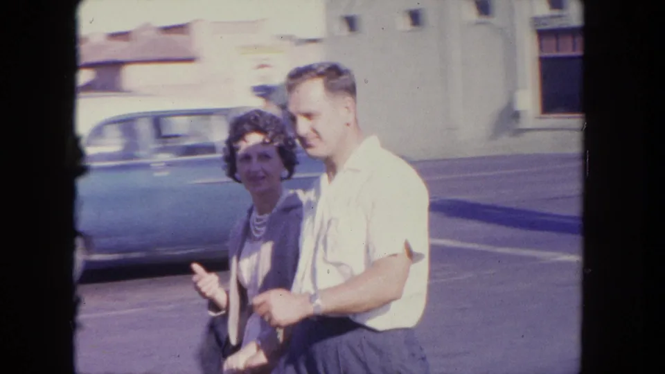 In Tijuana, Mexico, a woman and man walk past the camera on a sunny street with mid-century cars behind them.
