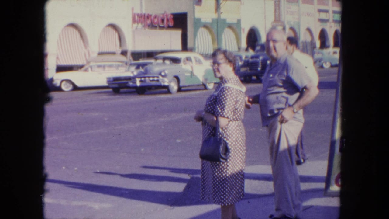 Older couple standing near a street with parked cars and arched storefronts behind them.