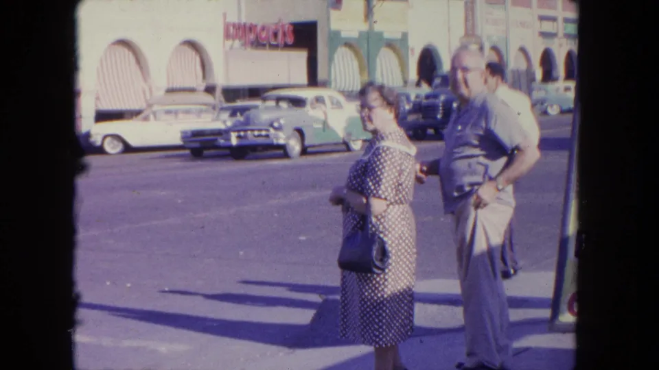 An older couple stands near a Tijuana street, with parked 1960s cars, arched storefronts, and an Imports sign in the background.