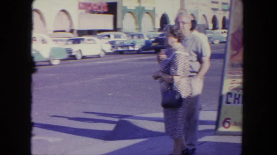 An older couple walks along a sunny Tijuana street, with classic cars, storefront arches, and part of a poster visible nearby.