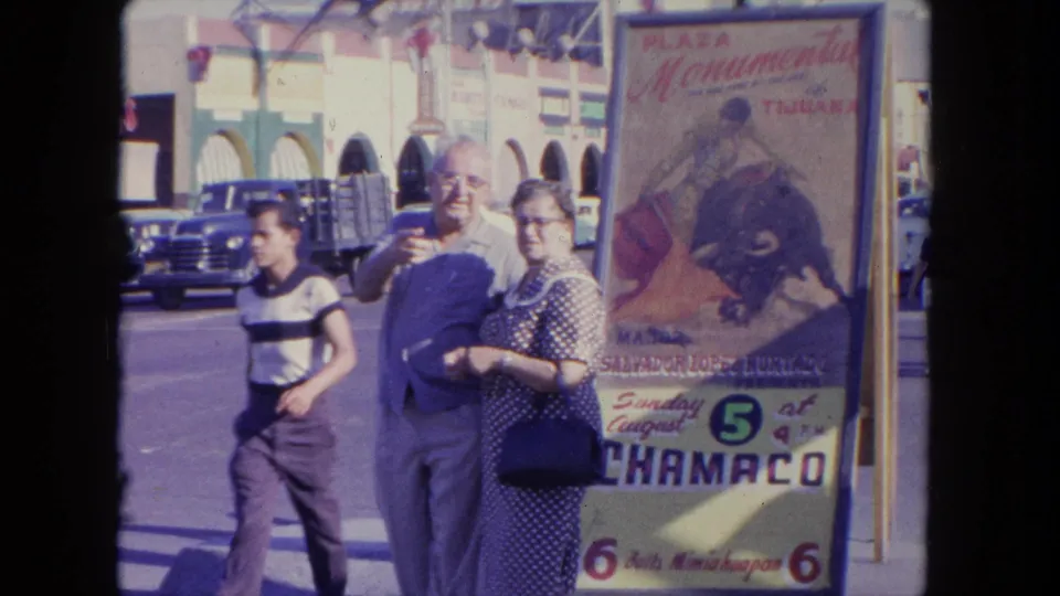 An older couple and a boy walk beside a Plaza Monumental Tijuana bullfight poster, with cars and arched storefronts across the street.
