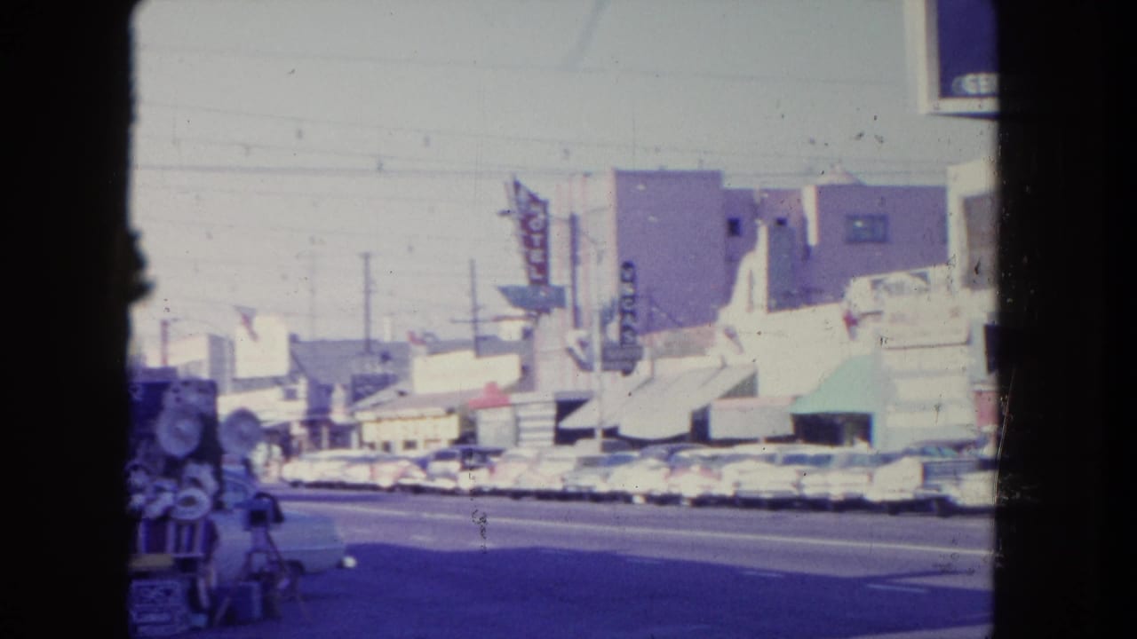 Sunlit commercial street with parked cars, storefront awnings, motel signage, and a vendor cart at the edge of frame.