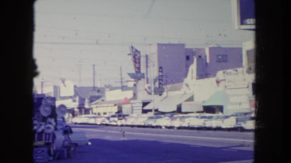 1962 Tijuana street view with mid-century cars parked along storefronts, vertical motel signage, awnings, and a vendor cart partly visible in front.