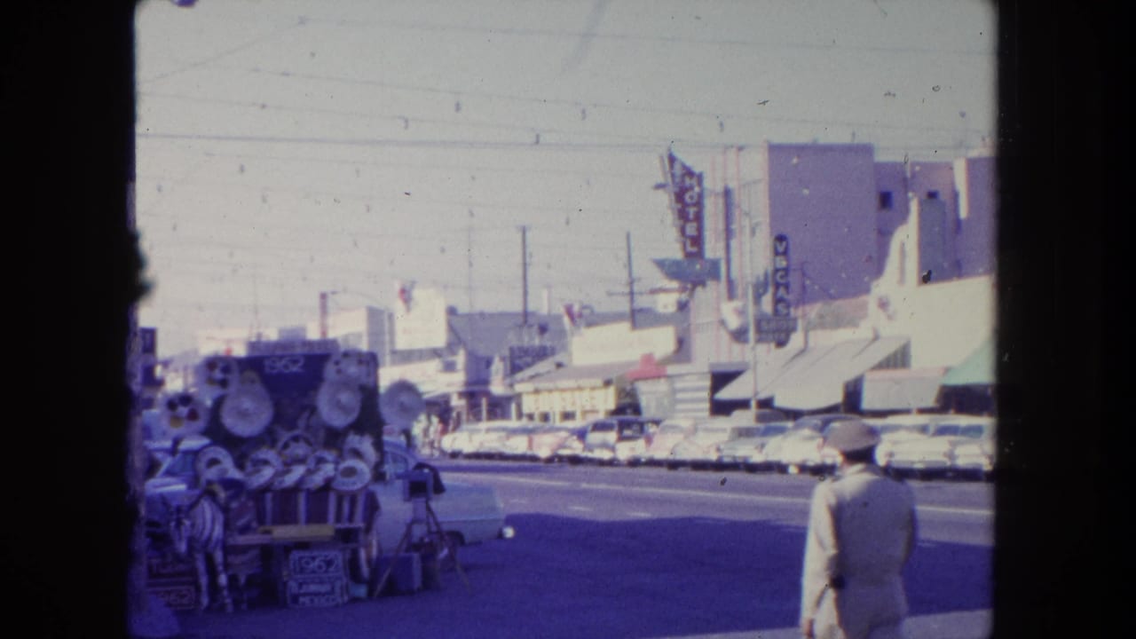In a 1962 Tijuana street scene, a pedestrian passes a vendor cart loaded with round metal parts near parked cars and visible motel signage.