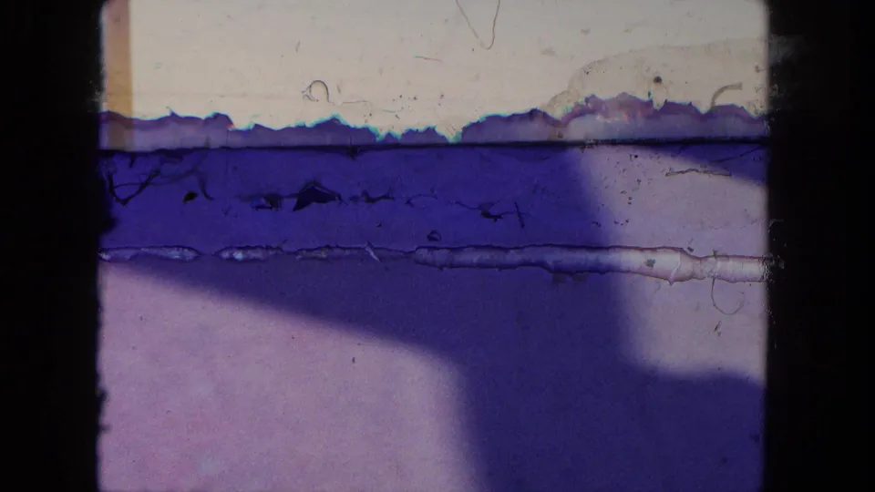 A heavily degraded 8mm frame from the Tijuana street clip shows blue-purple staining, scratches, and partial exposure with minimal readable street detail.