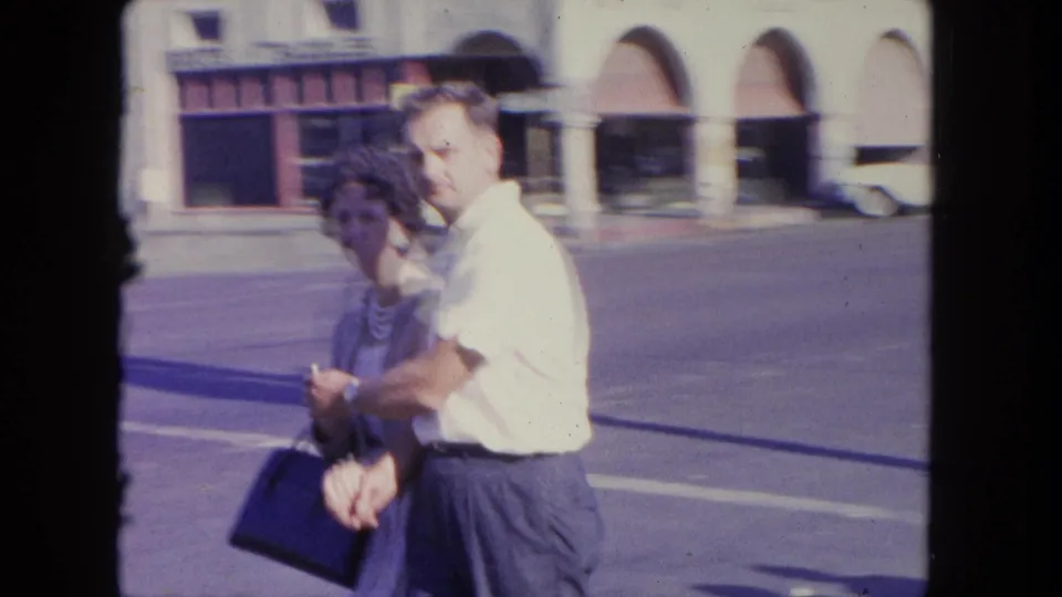A couple crosses a sunny Tijuana street in 1962, with the man looking toward the camera and arched storefronts blurred behind them.