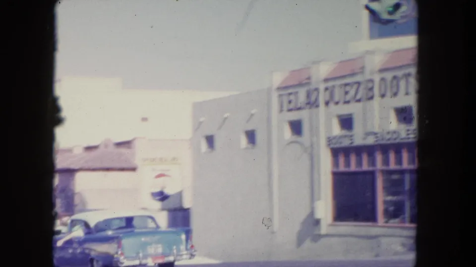1962 Tijuana street view with storefront signs, a parked blue sedan, and dark 8mm frame borders.