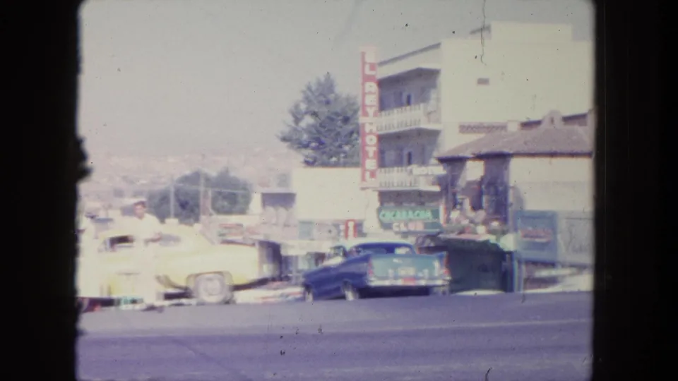 1962 Tijuana street scene with an El Rey Hotel sign, parked classic cars, pedestrians, and distant hillside.