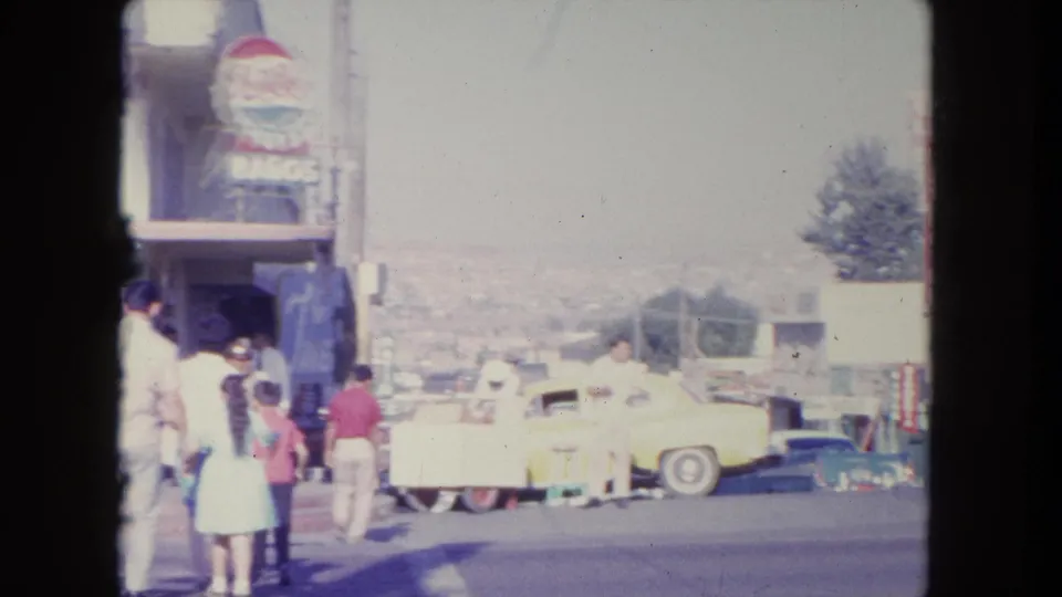 1962 Tijuana sidewalk scene with pedestrians, a vertical sign, a pale yellow car, and hazy hillside beyond the street.