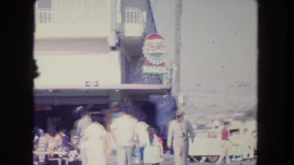 1962 Tijuana commercial street view with people near a storefront, a vertical sign, parked cars, and soft 8mm exposure.