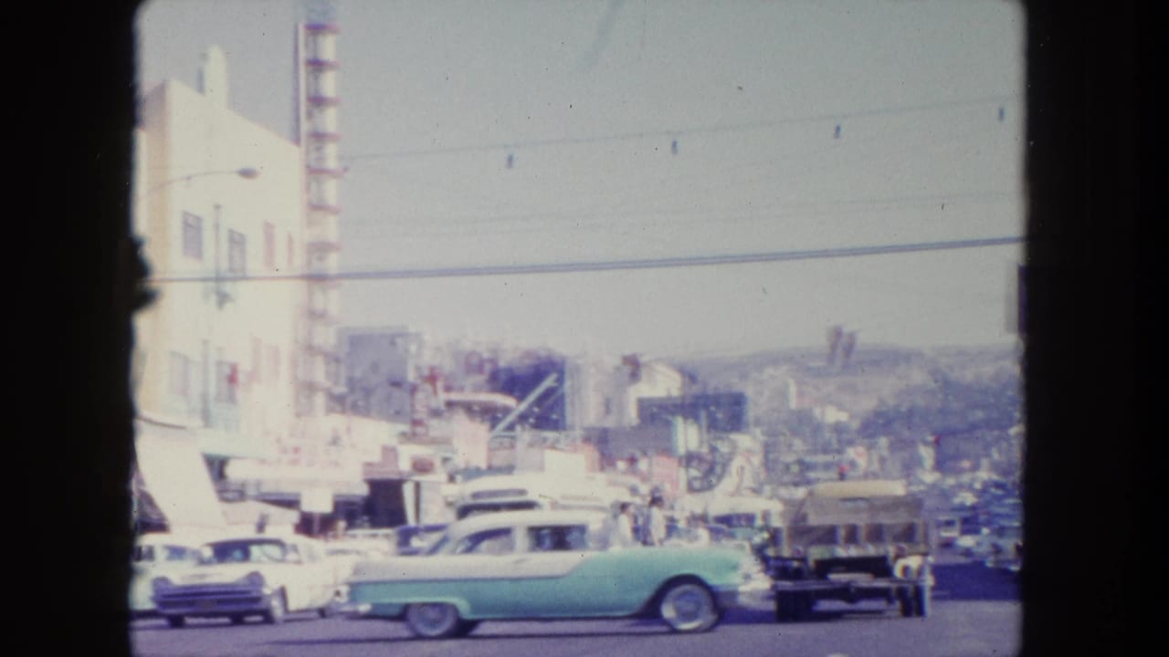 Busy street with classic cars, storefronts, utility wires, and hills in the distance.