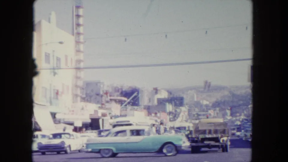 1962 Tijuana street view with early 1960s cars, pedestrians, storefronts, overhead wires, and hillside buildings beyond.