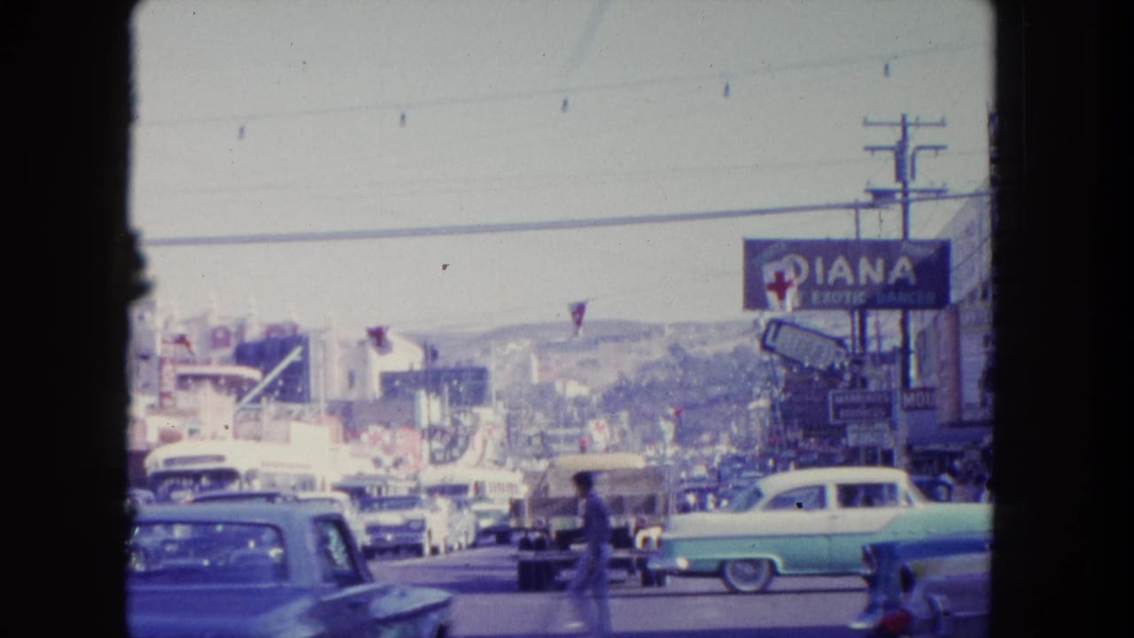 Street traffic passes storefront signs, parked cars, pedestrians, wires, and distant hills.