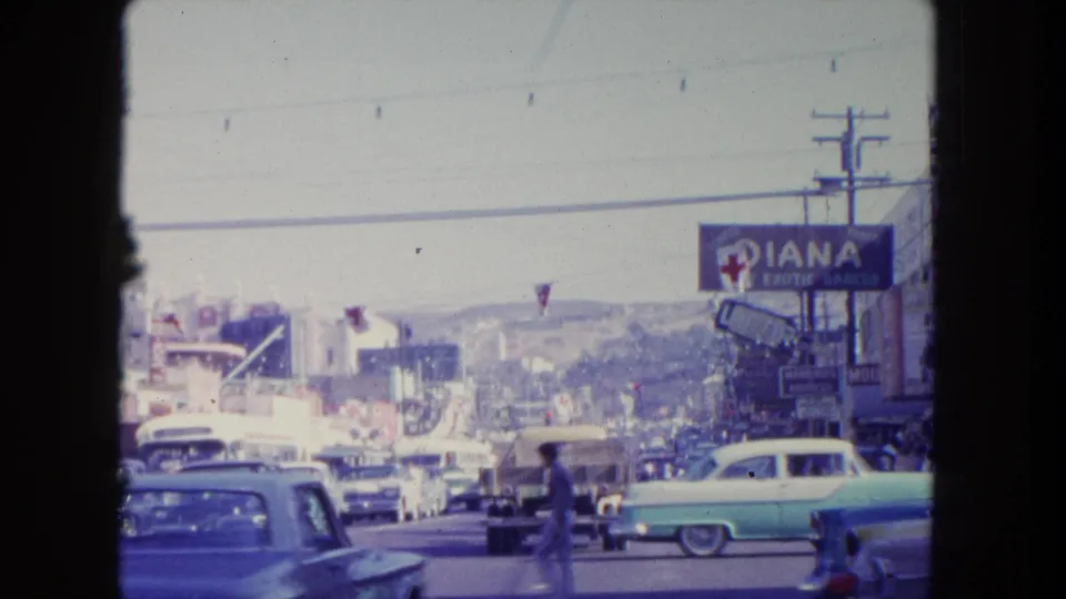 Moving 8mm view of Tijuana traffic with classic cars, pedestrians, a Diana sign, utility lines, and hillside cityscape.
