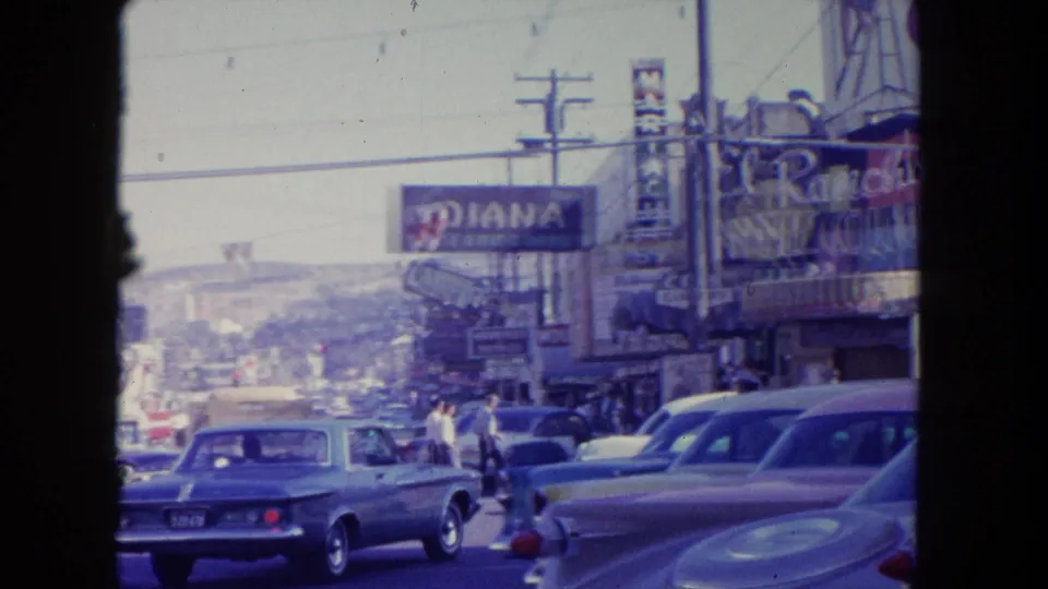 1962 Tijuana street scene with slow traffic, visible storefront signage, pedestrians, parked cars, and overhead utility lines.