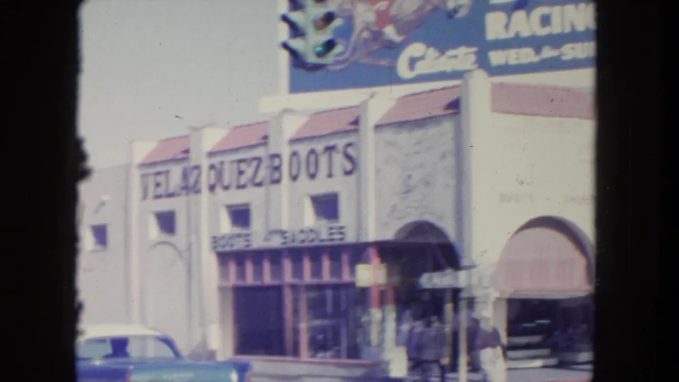 Tijuana storefront facade with large boot-shop lettering, street-level awnings, pedestrians, a parked car, and a racing billboard above.