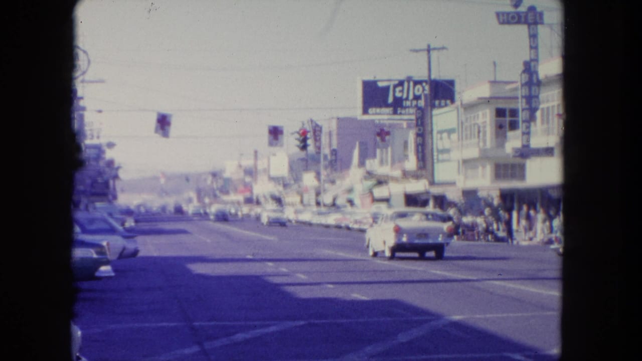 1962 Tijuana street view with classic cars moving past Tello's Import signage, traffic lights, flags, and pedestrians along storefronts.