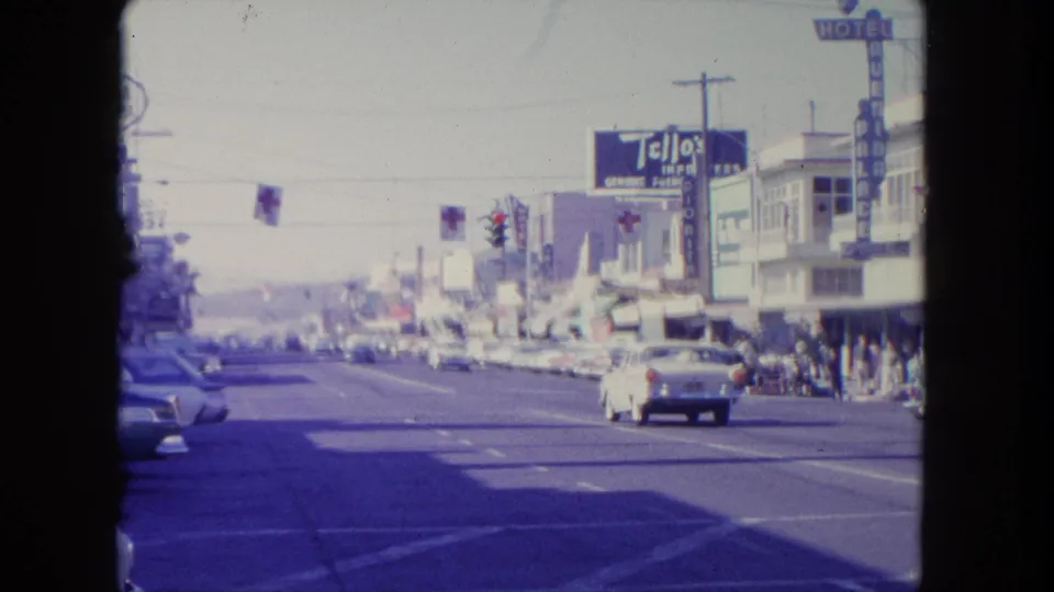 1962 Tijuana street view with classic cars moving past Tello's Import signage, traffic lights, flags, and pedestrians along storefronts.