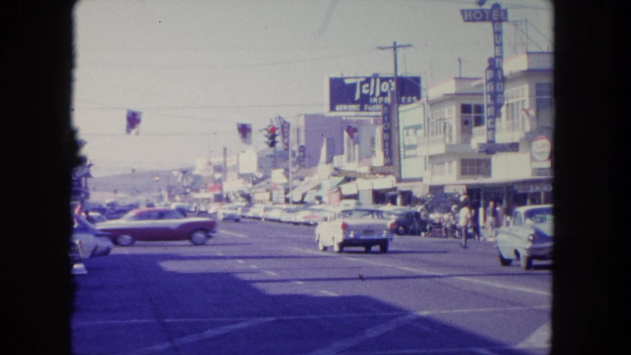 Red and white cars cross a broad street near storefronts, pedestrians, traffic lights, and hotel signage.