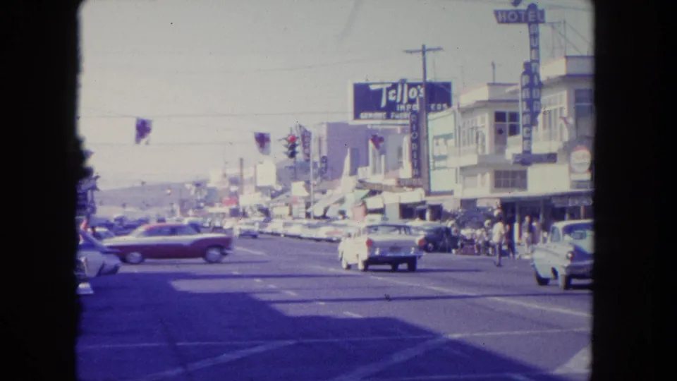 1962 Tijuana traffic scene with a red car crossing the street, white cars nearby, pedestrians at shops, and visible hotel and import signs.