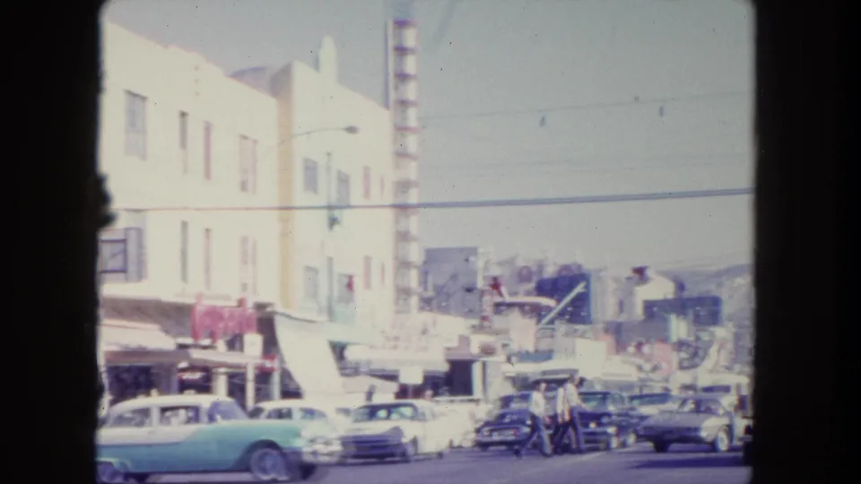 1962 Tijuana street view with green, white, and dark classic cars near shop awnings, pedestrians, vertical signage, and hills in the distance.