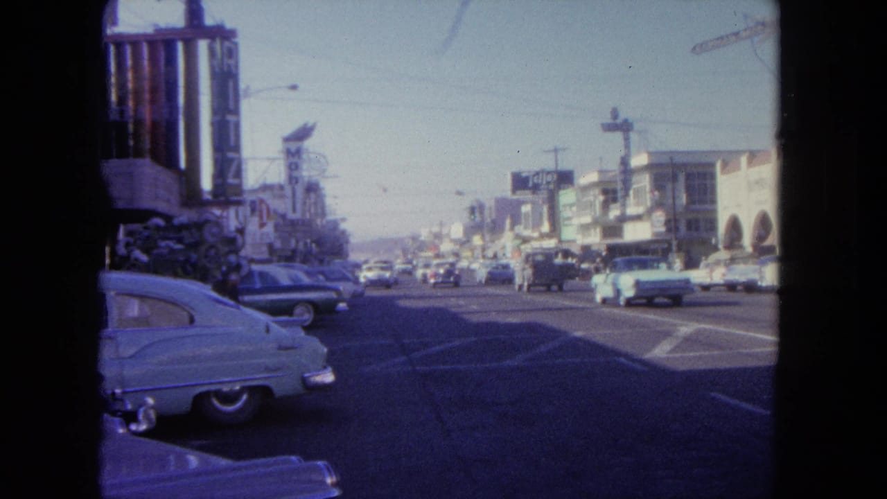 1962 8mm view of a Tijuana commercial street with classic cars, storefront signs, and traffic moving through a wide roadway.