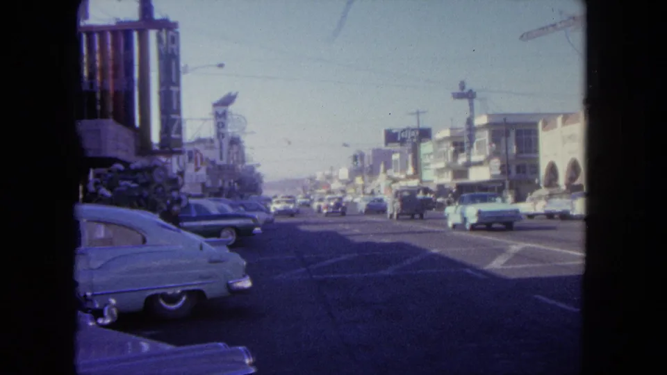 1962 8mm view of a Tijuana commercial street with classic cars, storefront signs, and traffic moving through a wide roadway.