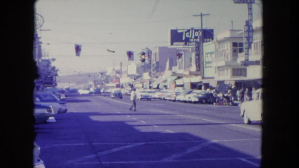 A man crosses at a Tijuana crosswalk in 1962 while classic cars and storefront signage fill the busy street scene.