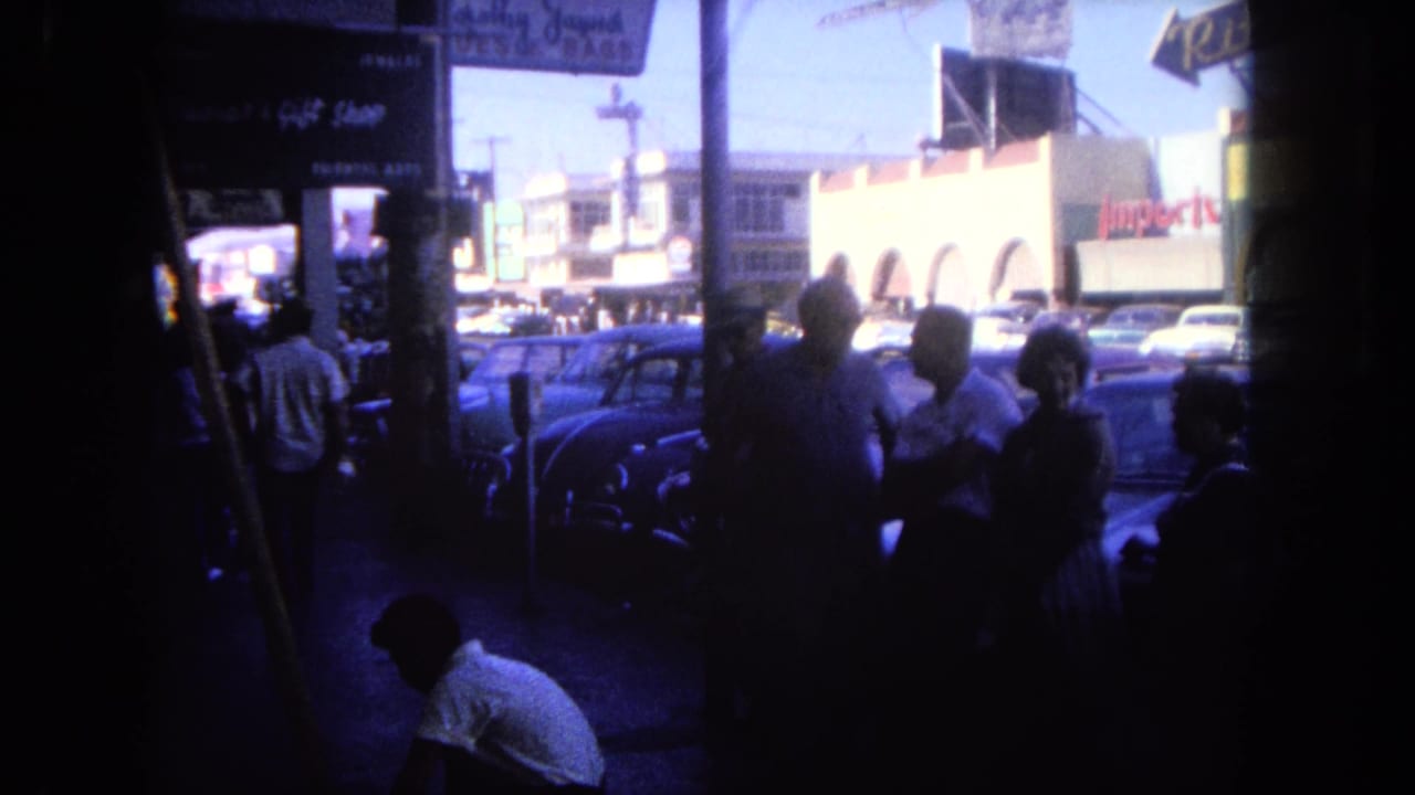 1962 Tijuana sidewalk scene with pedestrians in shade, storefront signs, and early 1960s cars parked along a sunny commercial street.