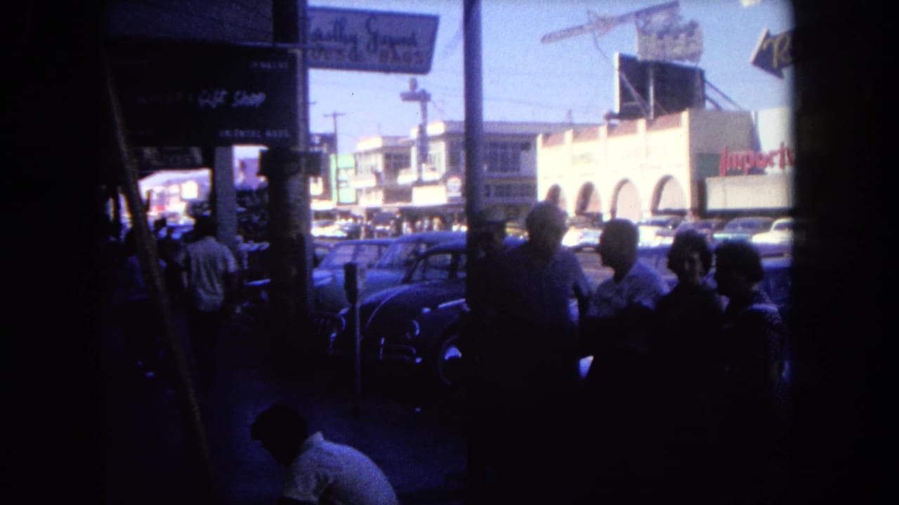 People gathered in shadow near storefronts with parked cars and commercial buildings across the street.