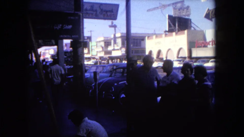 1962 Tijuana street view from shade, showing a small sidewalk group, storefront signage, parked cars, and sunlit commercial buildings.