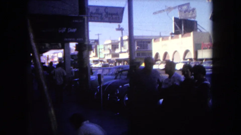 1962 Tijuana sidewalk footage showing pedestrians silhouetted in shade, storefront signs, and vintage cars along the commercial street.
